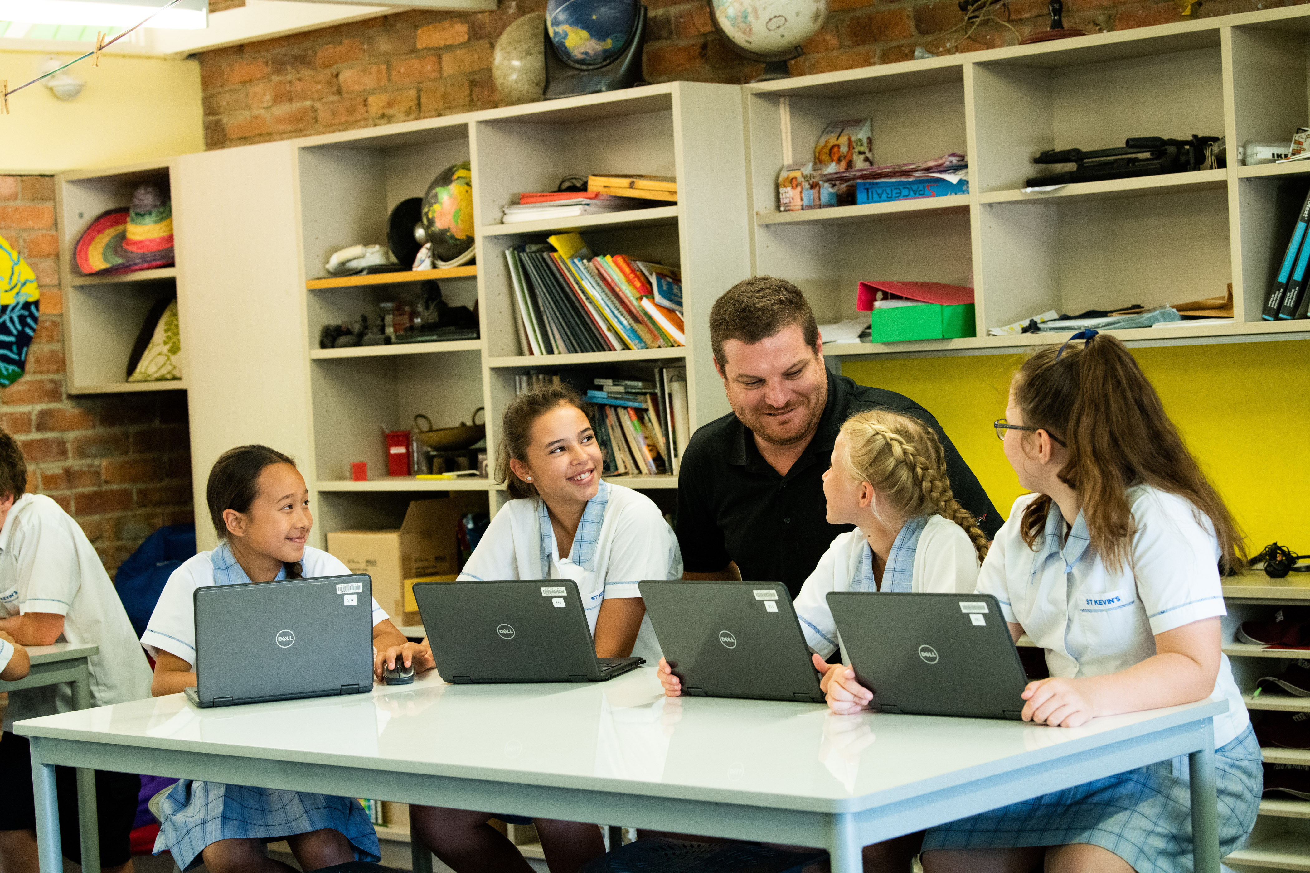 Four students are sitting at a desk and working on computers. They're teaching is kneeling behind them.